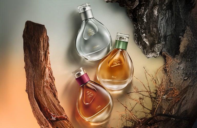 Foto de tres frascos de perfume Ekos de Natura, con forma de gota, con líquidos en tonos gris, ámbar y marrón, dispuestos sobre un fondo que simula madera y raíces.